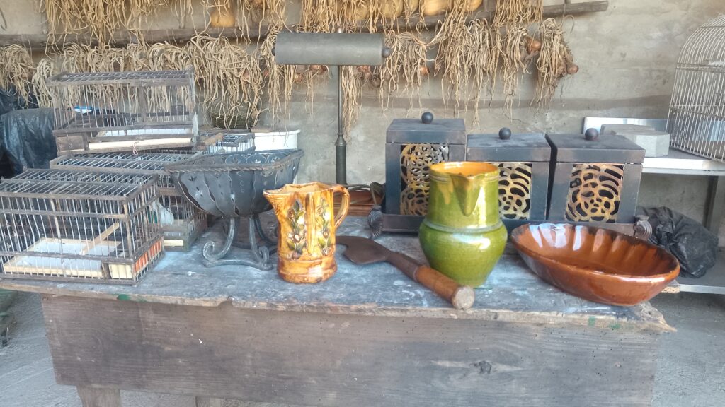 Sur une table sont présentés plusieurs objets d'art populaire, notamment 2 carafes en poteries (une verte et une marron), une coupe à fruits en fer, des cages à oiseaux fabriquées en bois et barreaux de fer, des boîtes de style chinois, un plat en poterie marron ainsi qu'une lampe de bureau en fer