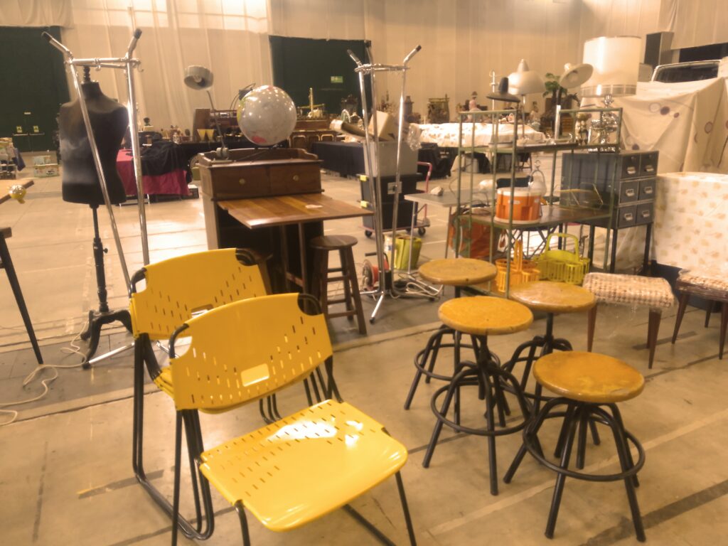 En premier plan, des chaises "Dallas" jaunes en fer.
En arrière plan, un meuble en bois exotique,  dont la porte sert de table et deux tabourets qui se rangent dedans.
Sur le coté, il y a des tabourets à vis en bois et fer