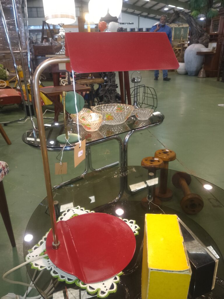 En premier plan, il y a une lampe de  rouge et cuivre ainsi qu'un reveil de marque "Jaz" , posés sur une table basse en verre et pied chromé.
En second plan, un service à salade de fruits en verre avec décors, des annéees 70, est posé sur une autre table en verre et pied chromé
